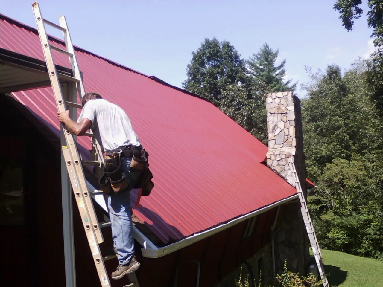 Roofer on a ladder beside a red metal roof and stone chimney — Soffit & Fascia Repair by Roofing Brothers in Kittery